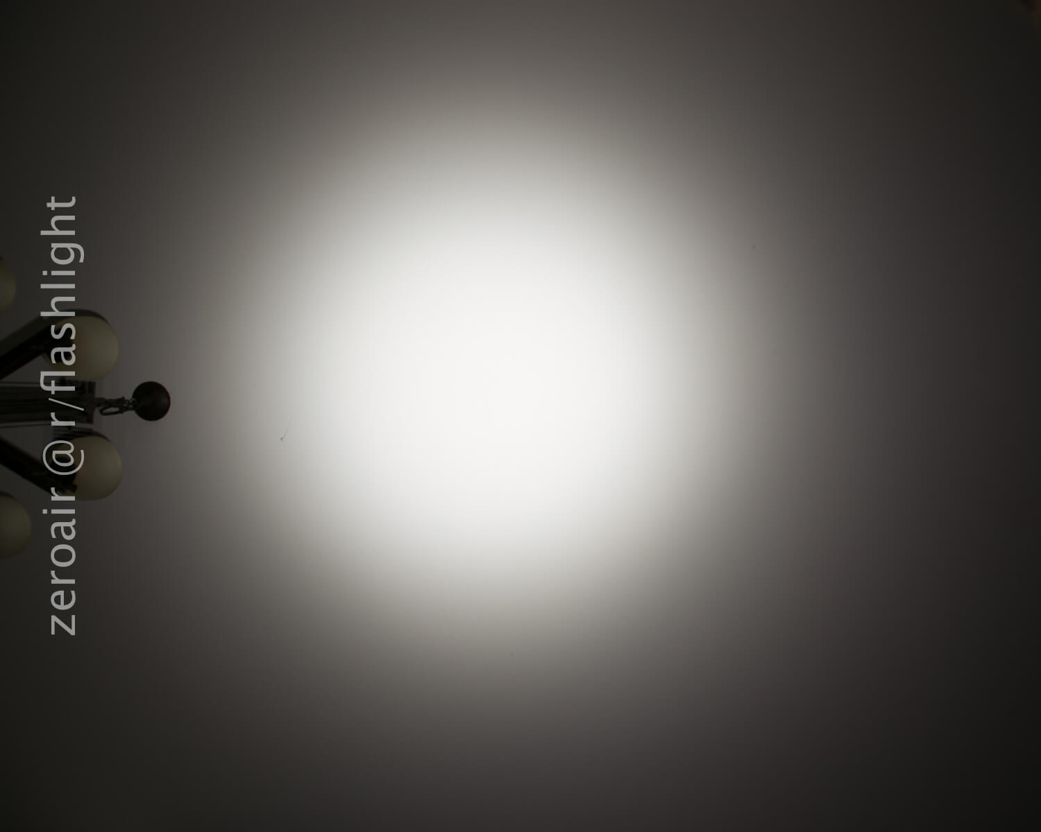A bright circular beam of light is projected onto a plain wall. In the upper left corner, part of a ceiling light fixture with multiple white globes is visible. Text along the left side reads zeroair @ r/flashlight.
