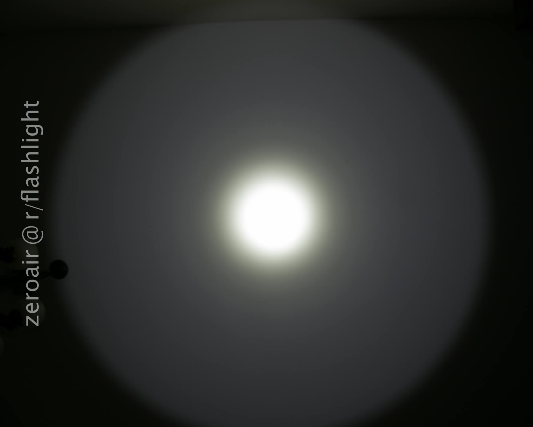 A circular beam of white light from a flashlight is projected onto a flat, dark surface, creating a bright center with a gradual fade toward the edges. The watermark zeroair @r/flashlight appears on the left side.
