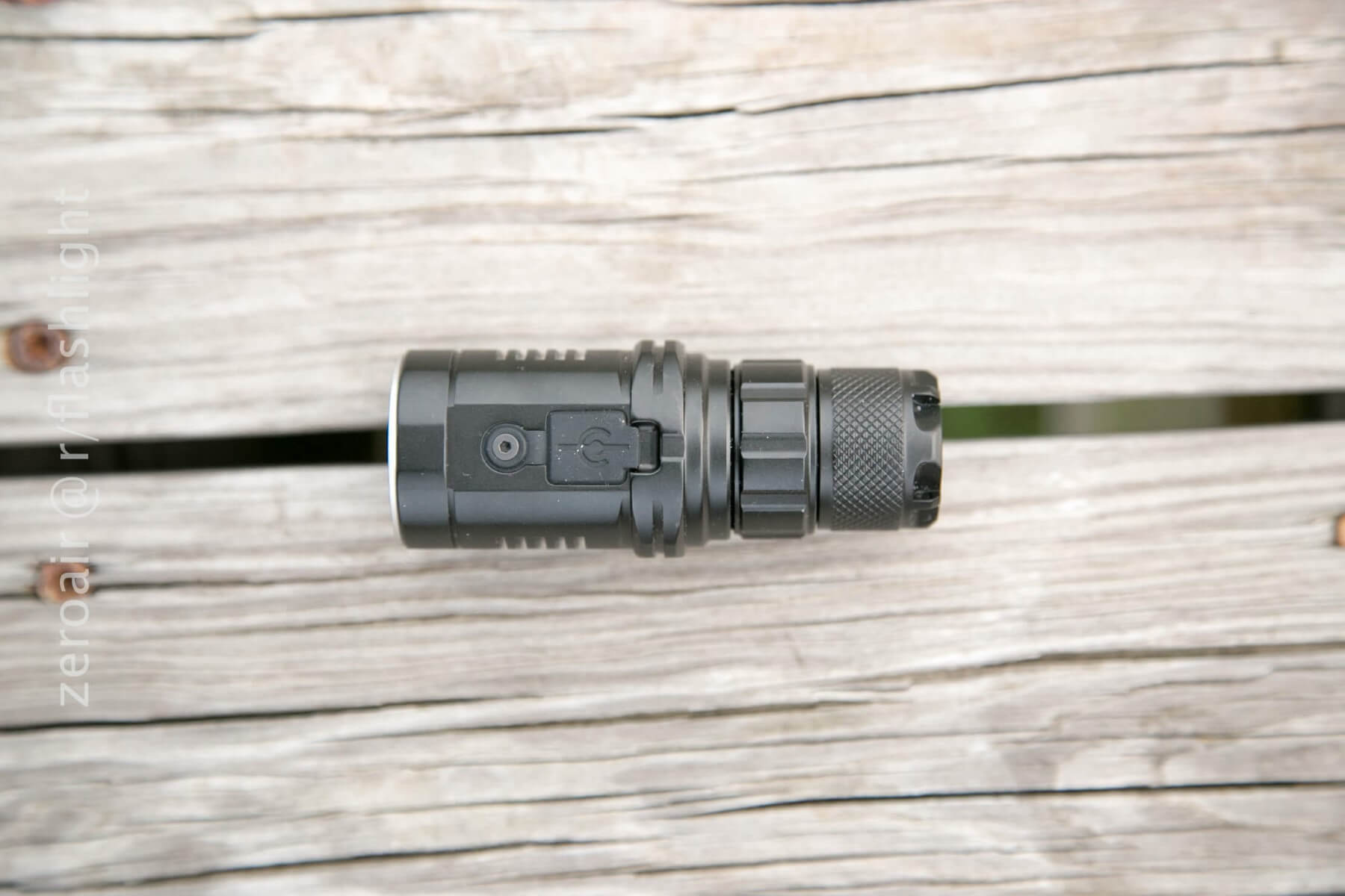 A small black Imalent DN12 flashlight lies horizontally on weathered wooden planks. The flashlight features a textured grip and a side button.