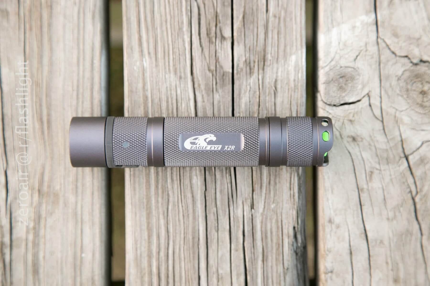 A cylindrical gray flashlight labeled Eagle Eye X2R rests horizontally on weathered wooden planks. The flashlight has textured grips and a green button on the end.