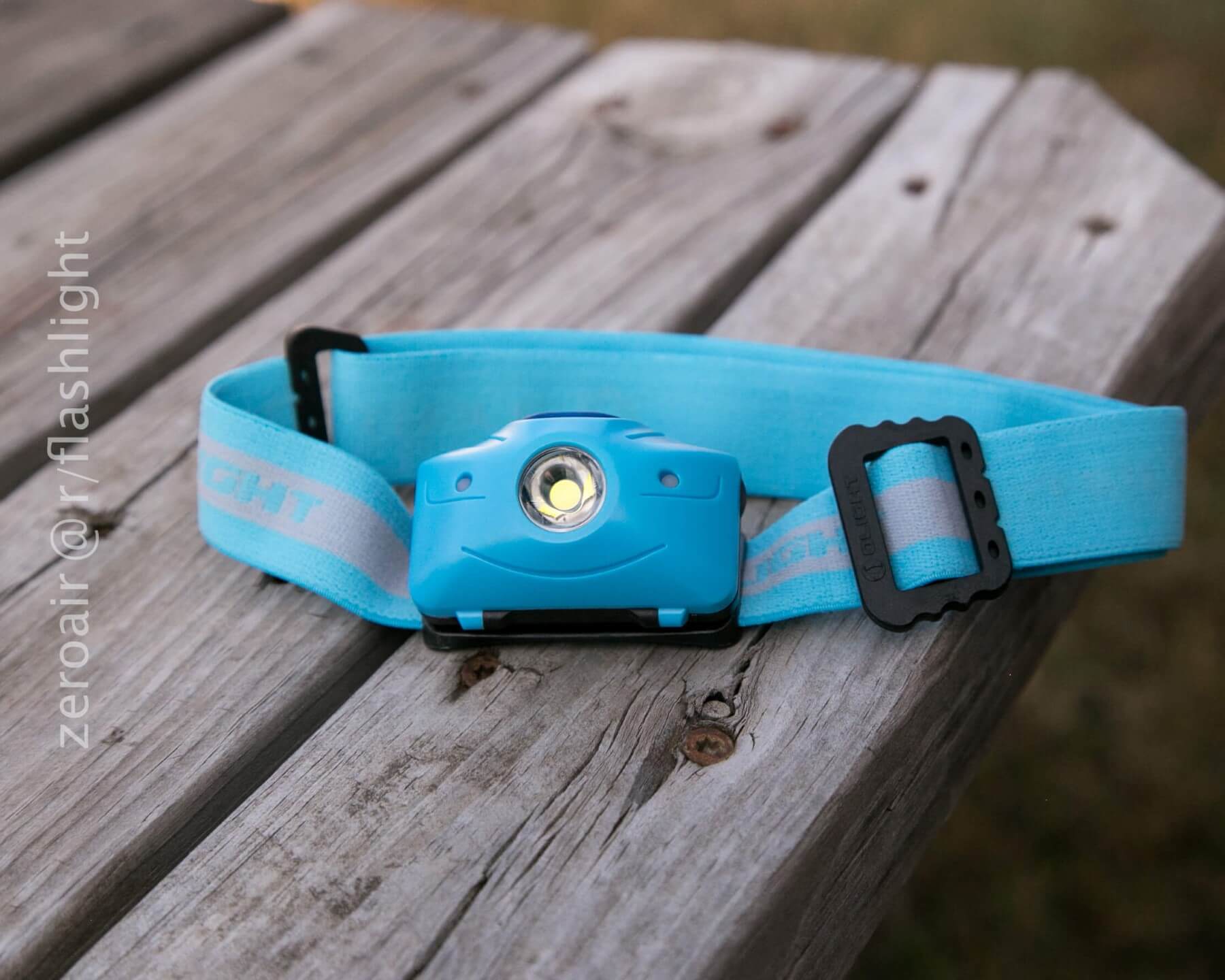 A blue and white headlamp rests on a weathered wooden surface outdoors, with the adjustable strap and LED light facing forward. The words zeroair/@/flashlight appear along the left side of the image.