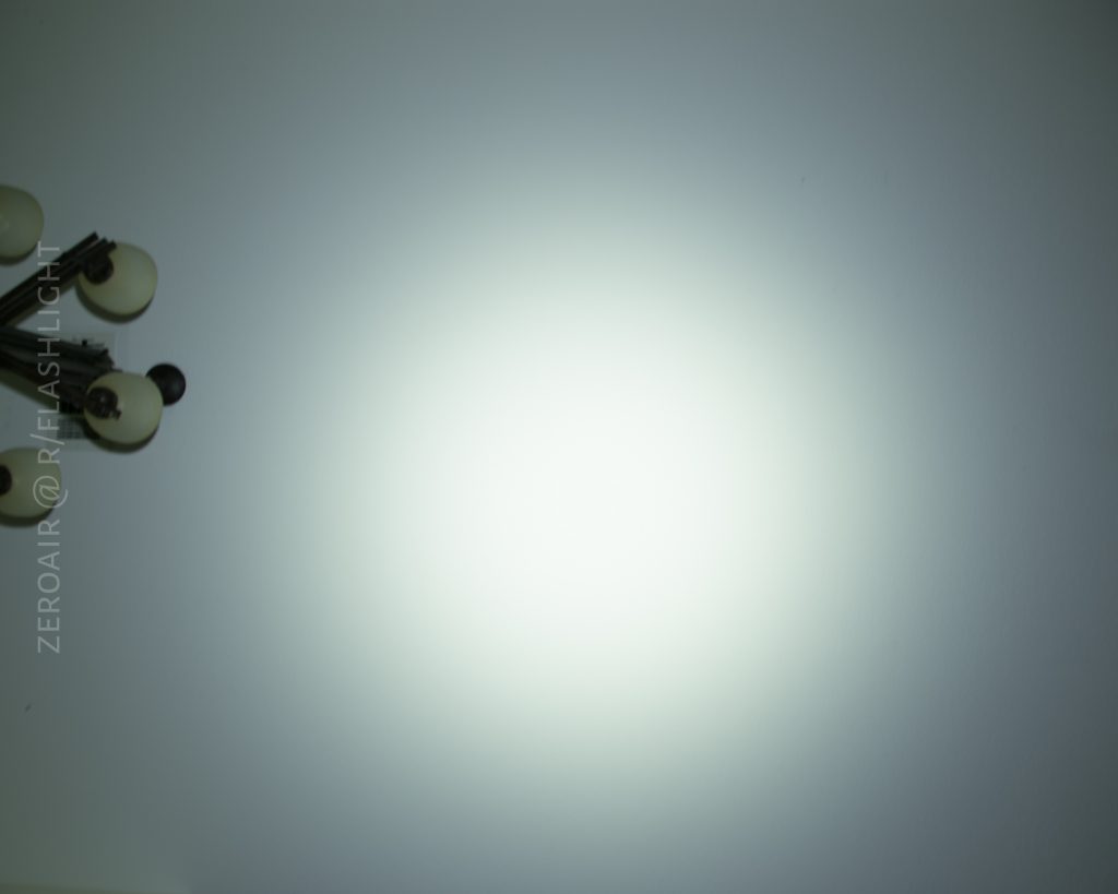 A bright circular flashlight beam is projected onto a plain white wall. In the upper left corner, part of a light fixture with several bulbs is visible. The text ZEROAIR @ ZEROAIR.ORG is positioned vertically on the left side.