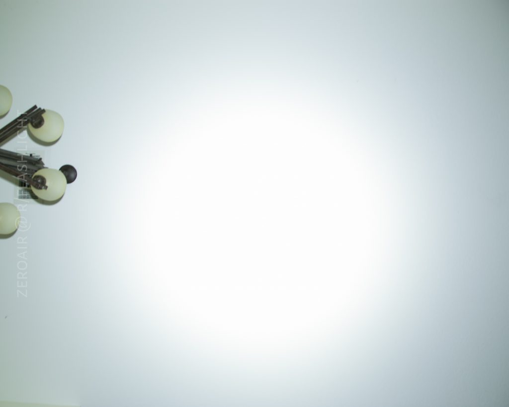 A ceiling with a bright circular light spot in the center, likely from a flashlight, and part of a multi-bulb ceiling light fixture visible in the upper left corner. The rest of the ceiling is plain and white.