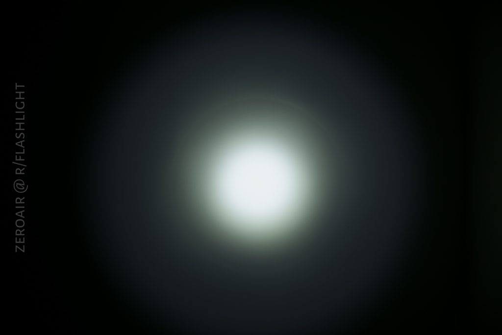A circular beam of white light shines on a dark background, creating a bright center that gradually fades to darker edges. Text along the left side reads ZEROAIR @ R/FLASHLIGHT.