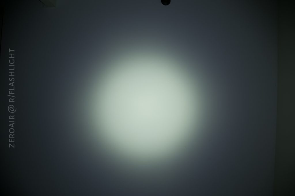A circular white beam of light is projected onto a gray wall in a dark room. The light appears brightest at the center and gradually fades toward the edges. Text along the left side reads ZEROAIR @ R/FLASHLIGHT.