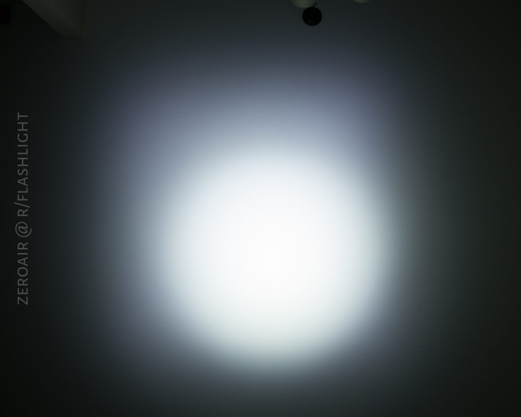 A circular beam of white light from the Olight S1Rv2 is projected onto a dark surface, creating a bright center with a gradual fade to darkness. The text ZEROAIR @ R/FLASHLIGHT is vertically aligned on the left side.
