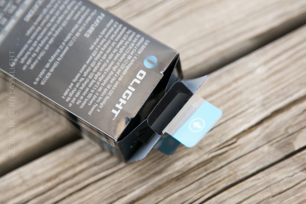 A close-up of an opened black Olight S1Rv2 product box with blue and black tabs sticking out, placed on a wooden surface. Text and branding are visible on the packaging.