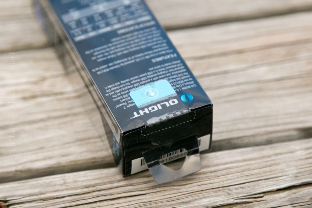 A close-up of a rectangular black box labeled OUGHT with a blue sticker seal partially opened, resting on a wooden surface. The packaging has small white text, barcodes visible on the flap, and resembles the style of an Olight S1Rv2 box.
