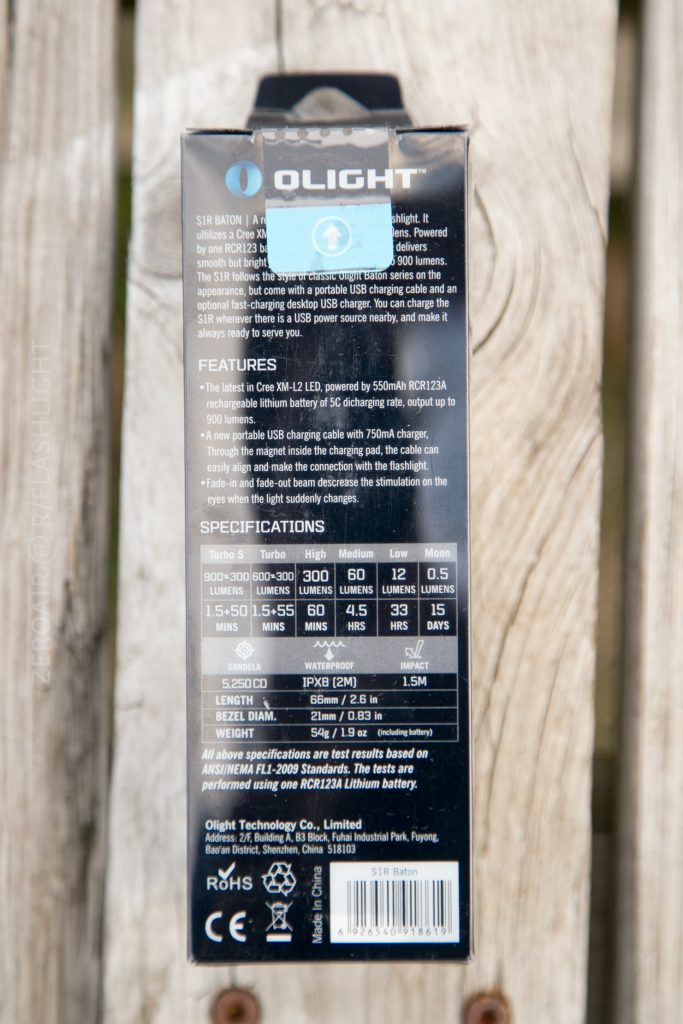 Back of an Olight S1Rv2 flashlight box showing features, specifications, and company information. Text displays performance details, barcode, certification logos, and a blue sticker with a battery symbol. Box rests on a wooden surface.