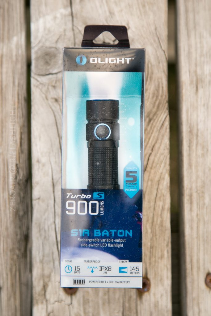 A boxed Olight S1Rv2 rechargeable LED flashlight is displayed upright on a wooden surface. The packaging highlights features such as 900 lumens, an IPX8 waterproof rating, and a 5-year warranty.
