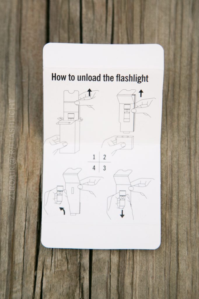 Instruction card on a wooden surface showing diagrams and text labeled How to unload the Olight S1Rv2 flashlight, illustrating step-by-step actions with arrows and hand drawings.