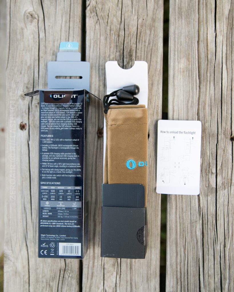A flashlight package is opened on a wooden surface, displaying the Olight S1Rv2 box, a tan carrying pouch with drawstring, and an instruction sheet with diagrams. The packaging highlights the features and specifications.