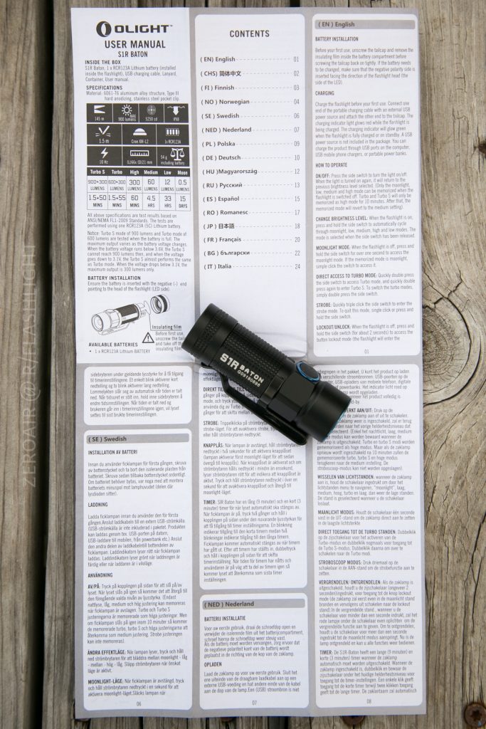 A black compact flashlight labeled Olight S1Rv2 Baton rests on top of an open user manual in multiple languages, all placed on a wooden surface. The manual contains safety information and specifications.