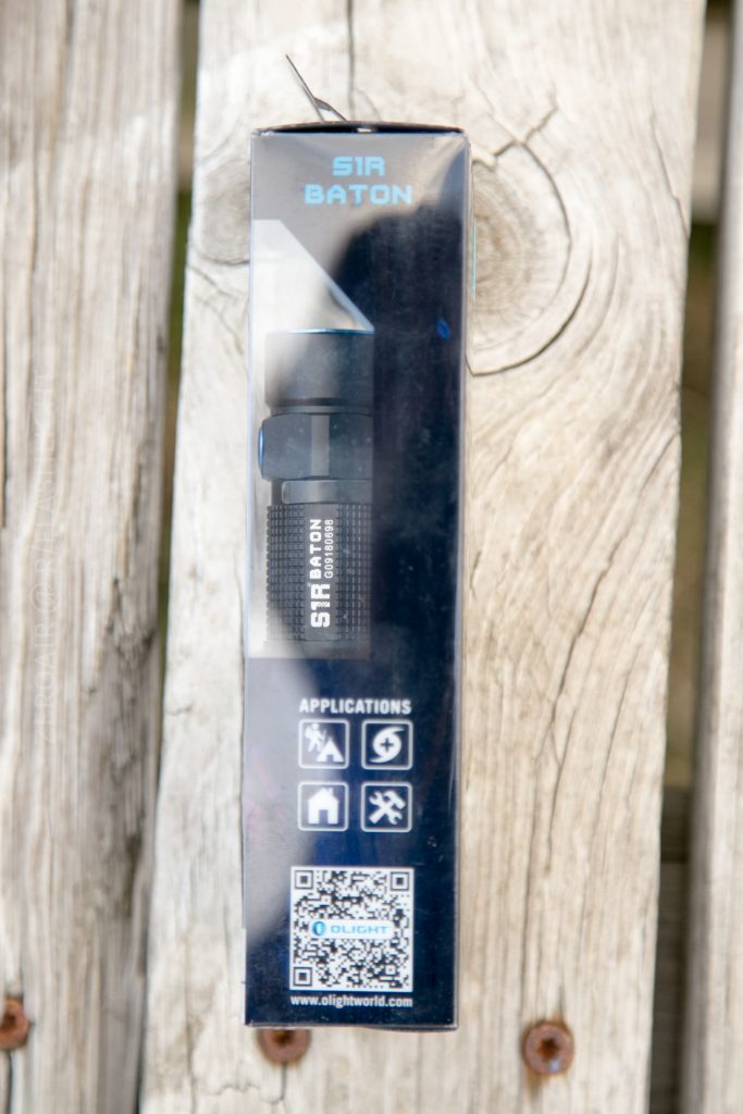 A boxed Olight S1Rv2 Baton flashlight stands upright on a wooden surface. The packaging displays the flashlight, four application icons, a QR code, and the brand's website.