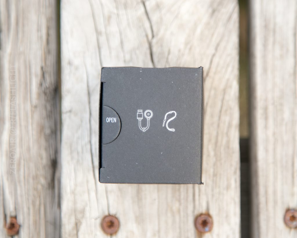A black box on a wooden surface, viewed from above, with white icons of a USB cable and an earpiece printed next to the word OPEN on one side, possibly packaging for the Olight S1Rv2.