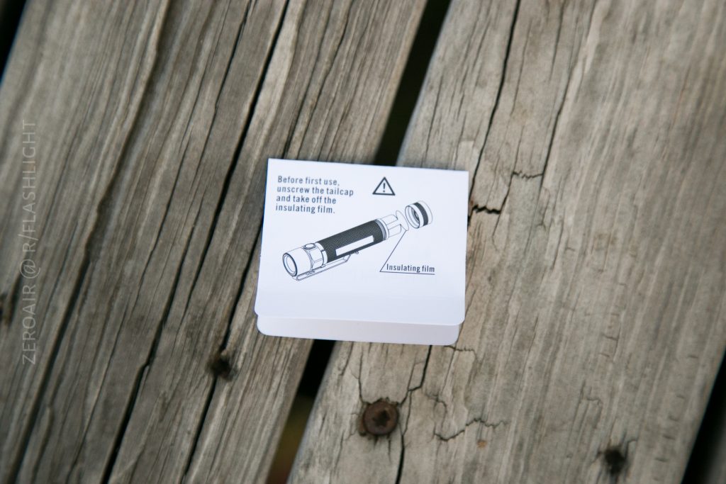 A small instruction card on a wooden surface shows a diagram of the Olight S1Rv2 flashlight and text advising to unscrew the tailcap and remove the insulating film before first use.