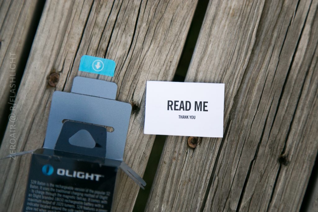 An open Olight S1Rv2 product box sits on a wooden surface next to a small white card that says READ ME THANK YOU in bold black letters.