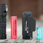 Four objects on a wooden surface: a black flashlight, a red 18650 lithium battery, a black Olight Valkyrie PL-2 flashlight, and a blue Olight 16340 battery. The background is blurred.