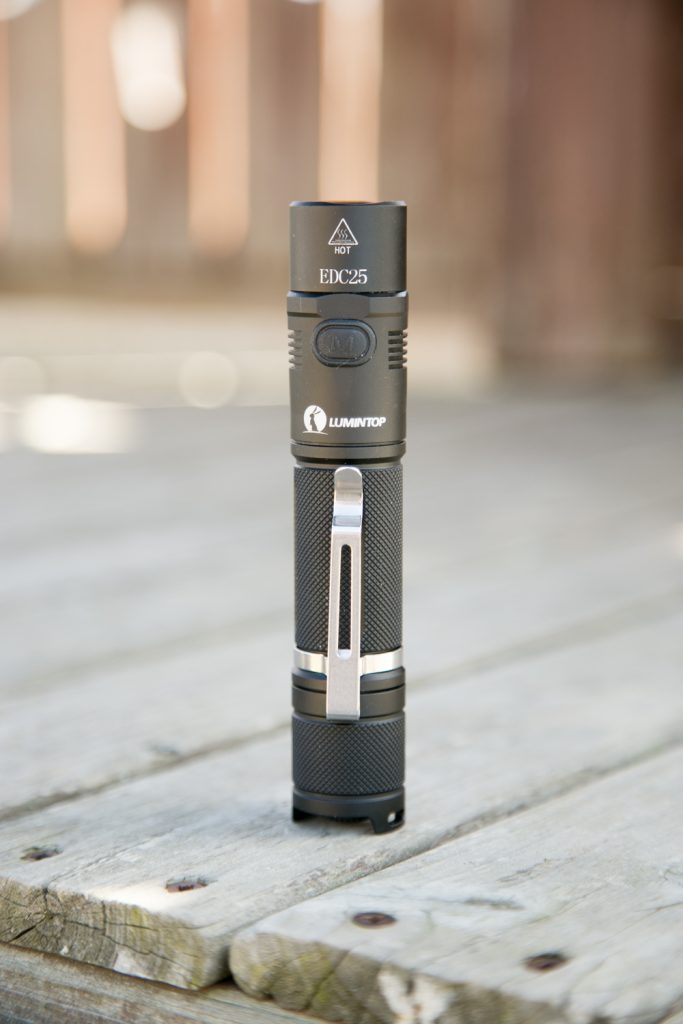 A black Lumintop EDC25 flashlight stands upright on a weathered wooden surface outdoors. The flashlight has a textured grip, metal clip, and visible brand markings. The background is blurred.