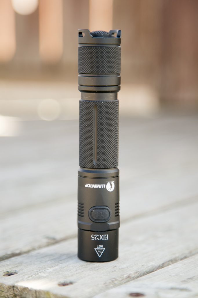 A black LUMINTOP EDC25 flashlight stands upright on a wooden surface. The textured grip, side button, and branding details are visible. The background is blurred.
