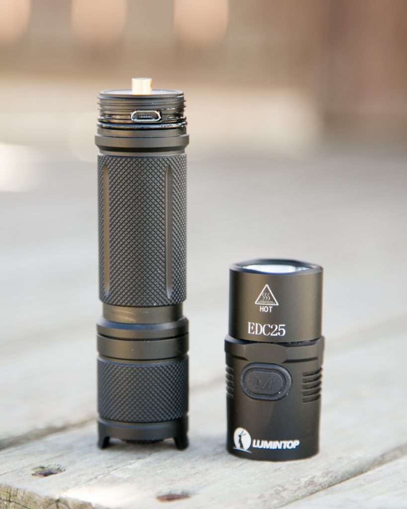 A black Lumintop EDC25 flashlight is shown disassembled into two parts on a wooden surface, revealing the battery compartment and metal threads.