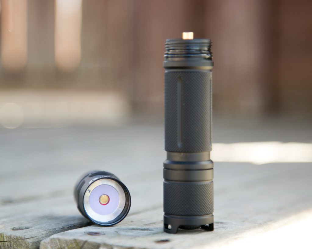 A black flashlight with a textured grip is separated into two parts, standing on a wooden surface outdoors. The head and body are detached, showing the LED bulb and battery compartment.