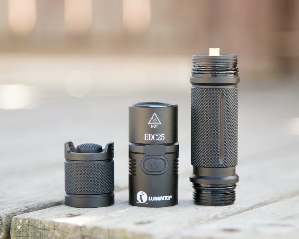 A black Lumintop EDC25 flashlight is disassembled into three parts and placed on a wooden surface. The parts include the tailcap, body tube, and head with visible threading and logos.