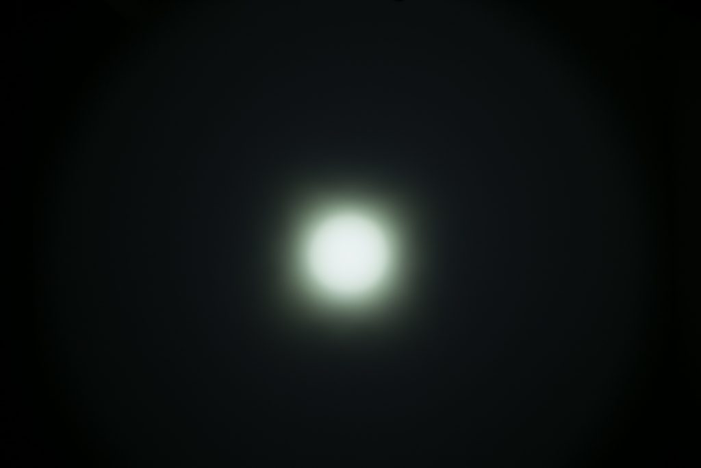 A bright, white circular light is centered against a dark background, surrounded by a soft, gradually fading glow. The edges of the light are blurred, creating a halo effect.