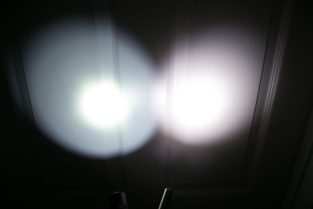 Two circular beams of light, one bluish and one white, are projected side by side onto a textured white surface with vertical grooves.