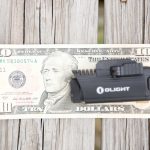 A black Olight flashlight is placed on top of a ten-dollar bill, both resting on a wooden surface with visible grain patterns.