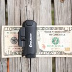 A black Olight flashlight rests horizontally on a ten-dollar bill placed on a wooden surface. The bill is unfolded and the flashlight covers the center.