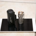 A black flashlight and a silver flashlight are standing upright on the screen of a white smartphone, which is placed on a tiled floor.