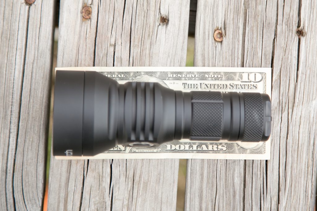 A black flashlight is placed horizontally on top of a ten-dollar bill, which rests on a weathered wooden surface. The flashlight is nearly as long as the bill.
