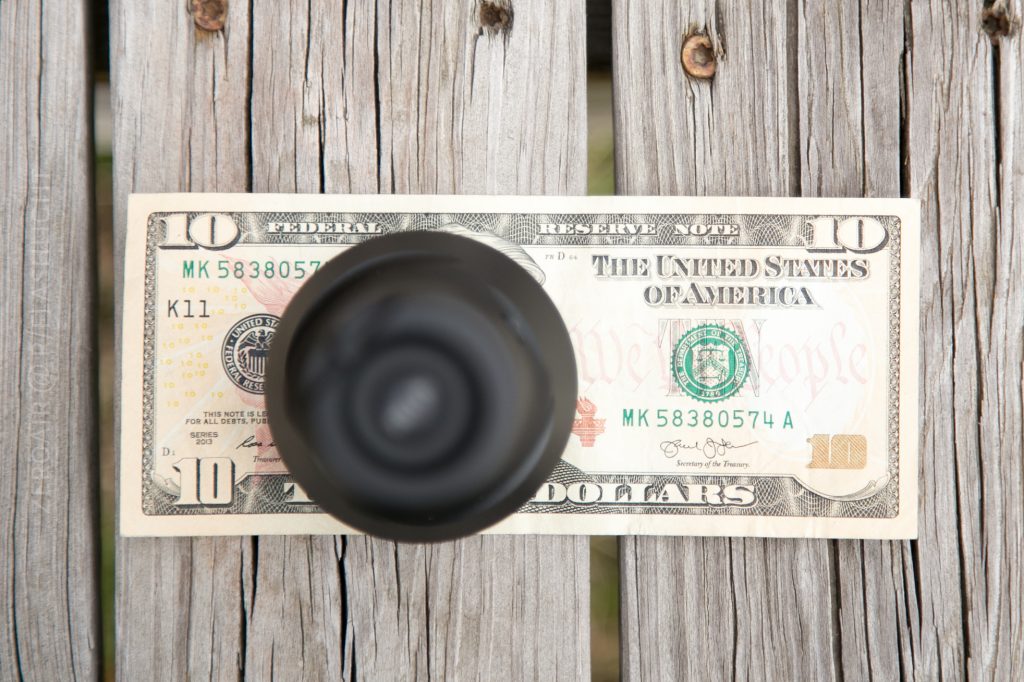 A ten-dollar bill is placed on a wooden surface with a black circular object positioned upright in the center, partially obscuring the bill.