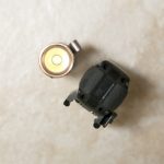 A top-down view of a flashlight with its lens facing up, next to a compact black device with textured grips, both placed on a beige surface.