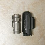 A small silver flashlight and a larger black tactical flashlight are placed side by side on a beige textured surface.