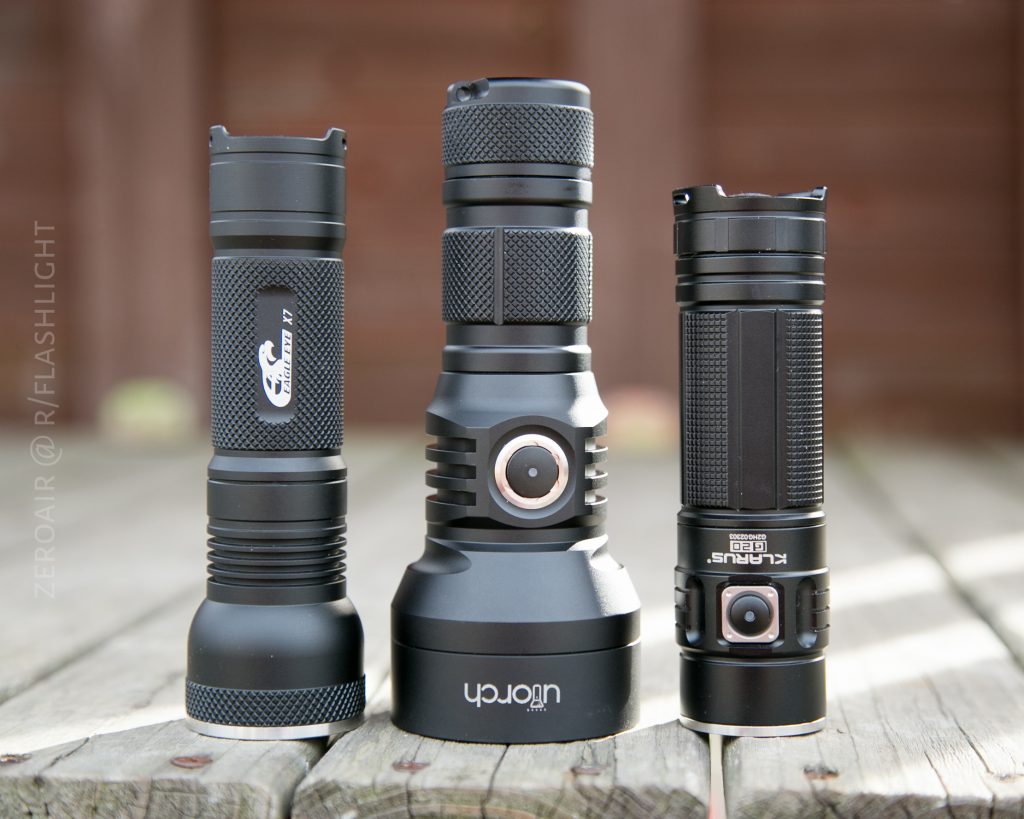 Three black flashlights of varying sizes and designs are standing upright on a wooden surface outdoors. The background is blurred, highlighting the flashlights in the foreground.