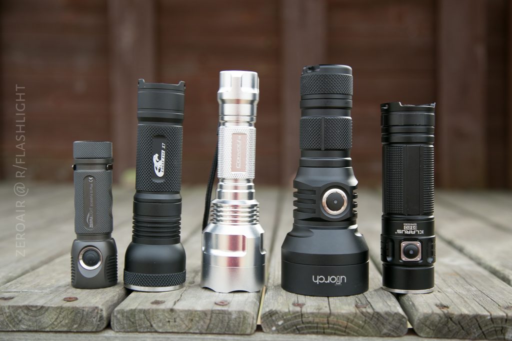 Five flashlights of different sizes and designs are standing upright on a wooden surface with a blurred wooden fence background. The flashlights vary in color, including black and metallic silver.