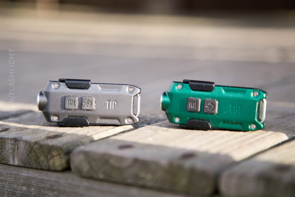 Two small flashlights, one gray and one green, are placed side by side on a wooden surface outdoors. Both have rectangular buttons and the word TIP embossed on their bodies.
