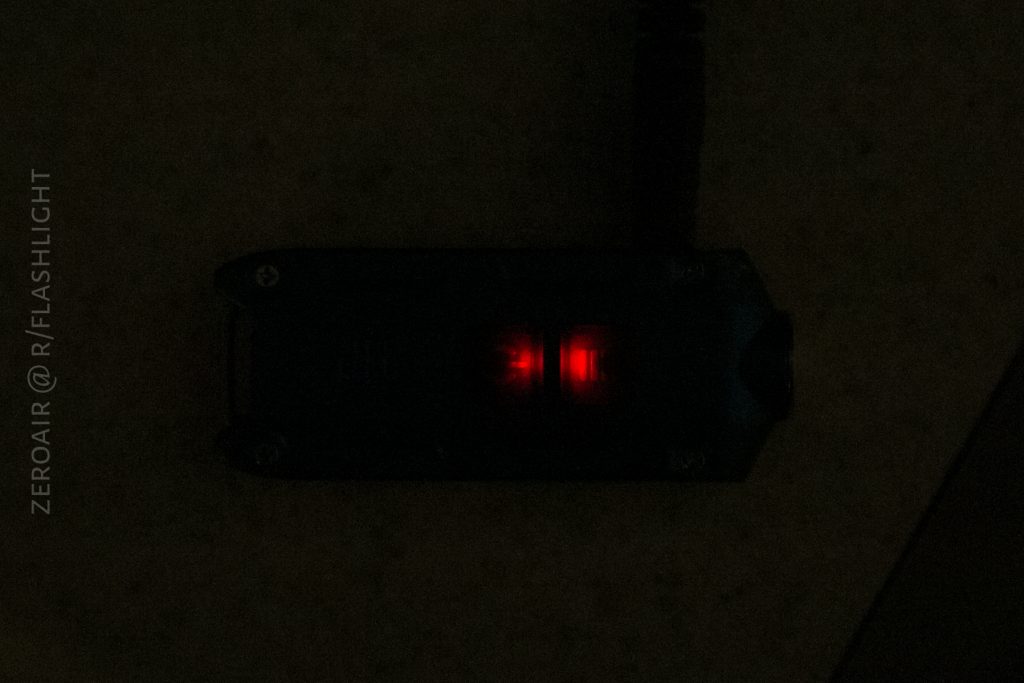A close-up of a dark charging device with two small red indicator lights illuminated. The background is dim, and a power cable is connected to the device. Text along the left side reads “ZEROAIR @ R/FLASHLIGHT.”