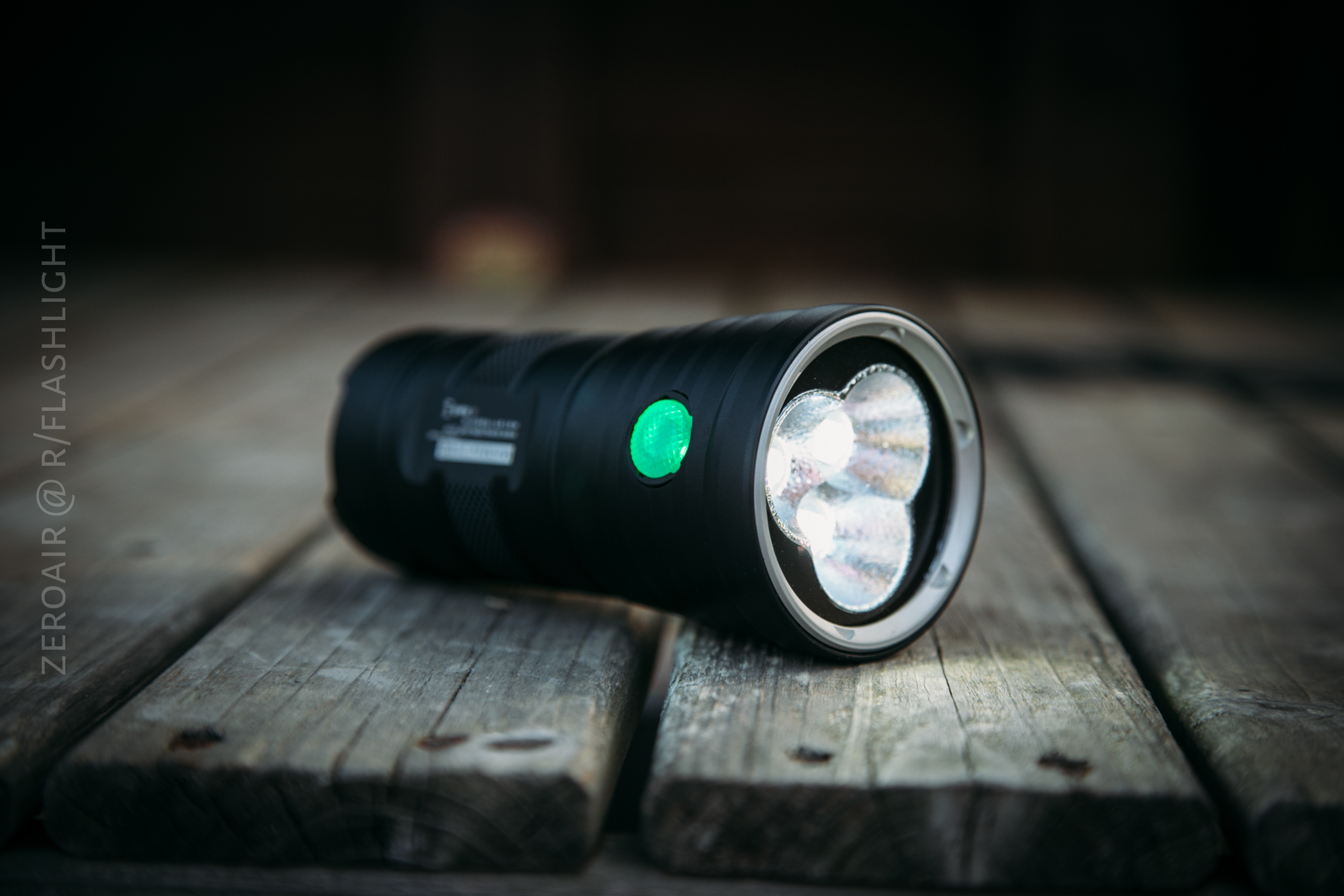 A black flashlight with a green power button is lying on a wooden surface, with its front facing slightly toward the camera and its light turned on.