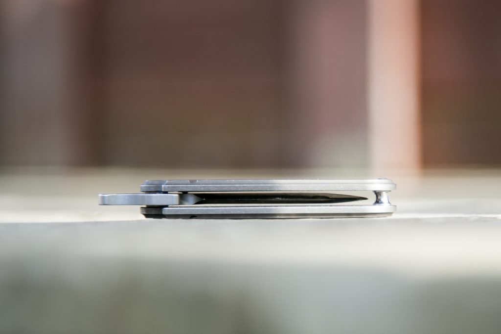 A closed gray multitool or folding pocket knife is lying flat on a light-colored surface, with an out-of-focus background in shades of brown and pink.