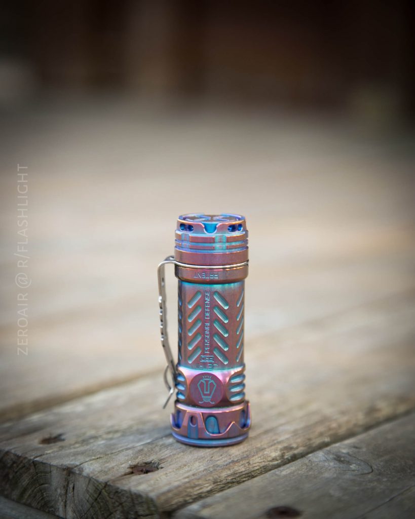A small, metallic flashlight with a blue and purple finish stands upright on a weathered wooden surface. The flashlight has a clip attached to its side and intricate patterns etched into its body.