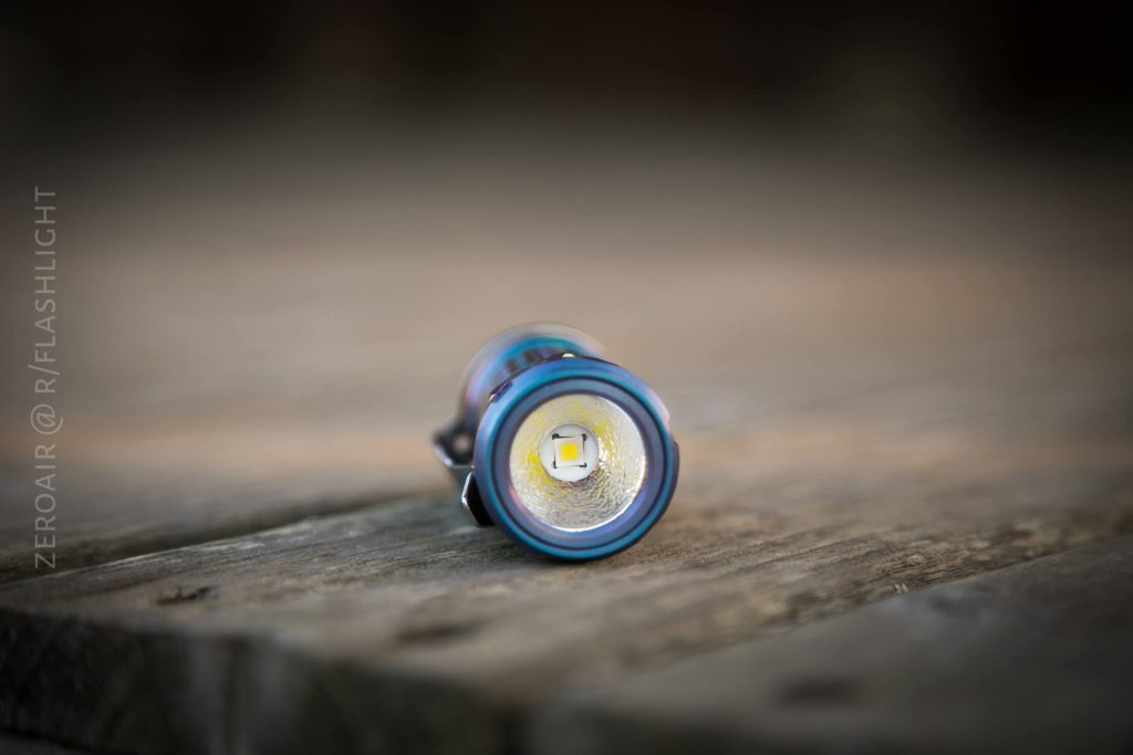 A small flashlight lies on its side on a wooden surface, with the lens and LED facing the camera. The background is out of focus. The flashlight appears metallic with a blue tint. Text is visible on the left side.