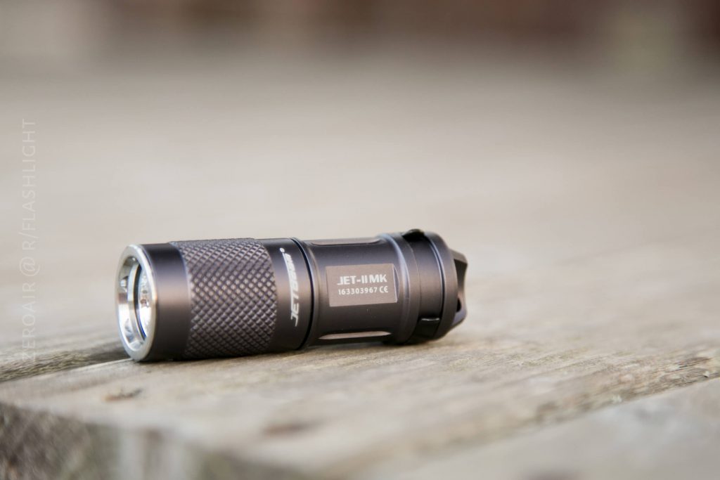 A small black JET-1 MK flashlight with textured grip rests on a wooden surface. The flashlight is positioned horizontally, and the brand name is clearly visible on its side.