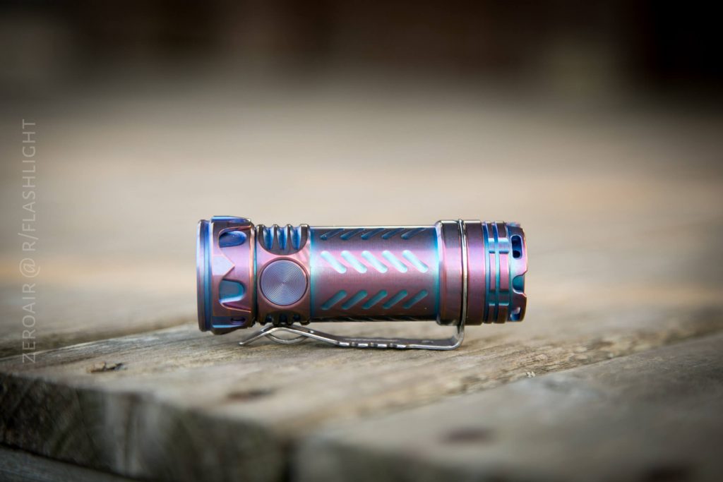 A metallic, multi-colored flashlight with blue and purple tones rests horizontally on a wooden surface. The item features grooves, a side button, and a metal clip. The background is blurred.