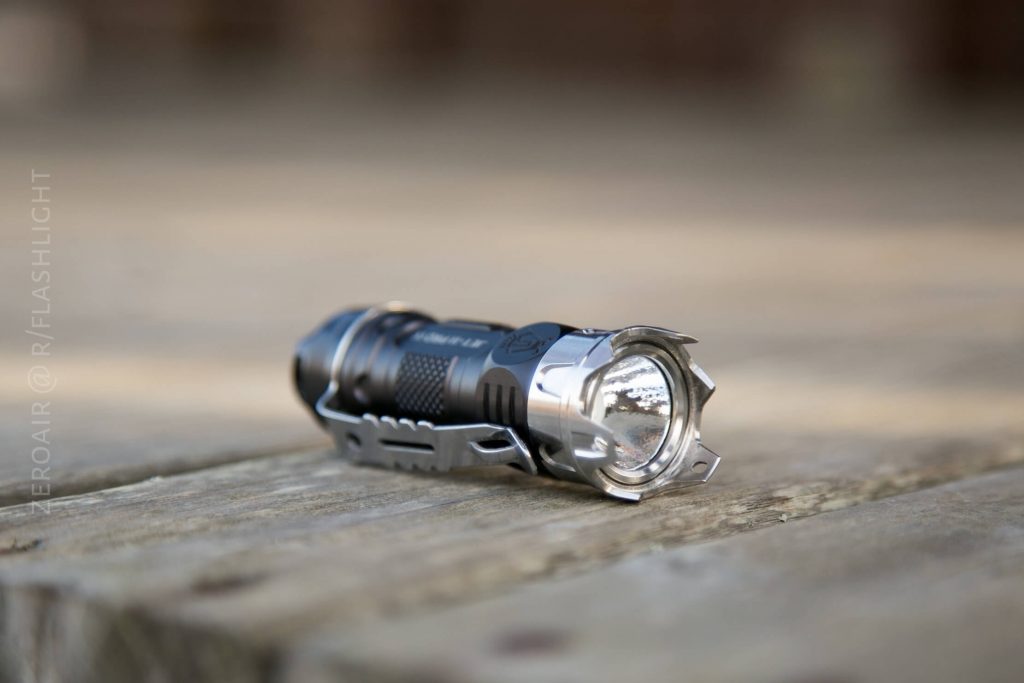 A compact, black and silver flashlight with a textured grip is lying on its side on a wooden surface, outdoors. The lens and metal bezel are clearly visible in the foreground.