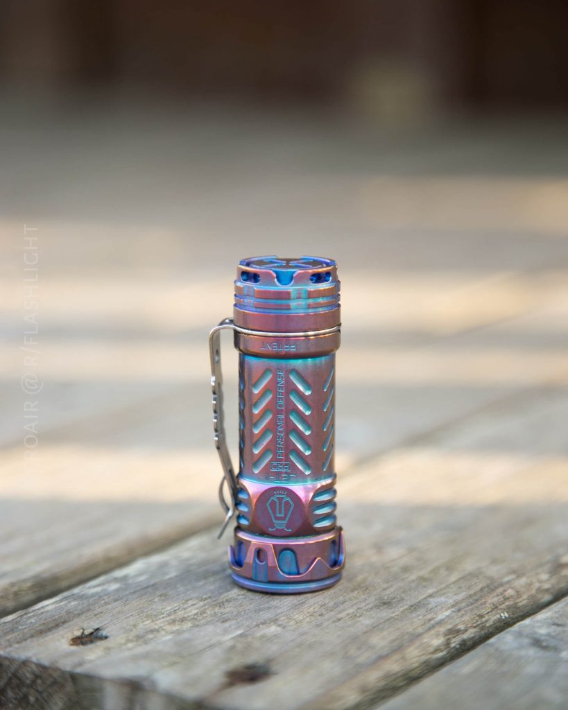 A metallic, multi-colored flashlight with textured grip and a clip stands upright on a wooden surface. The flashlight features blue, pink, and purple hues with a geometric pattern.
