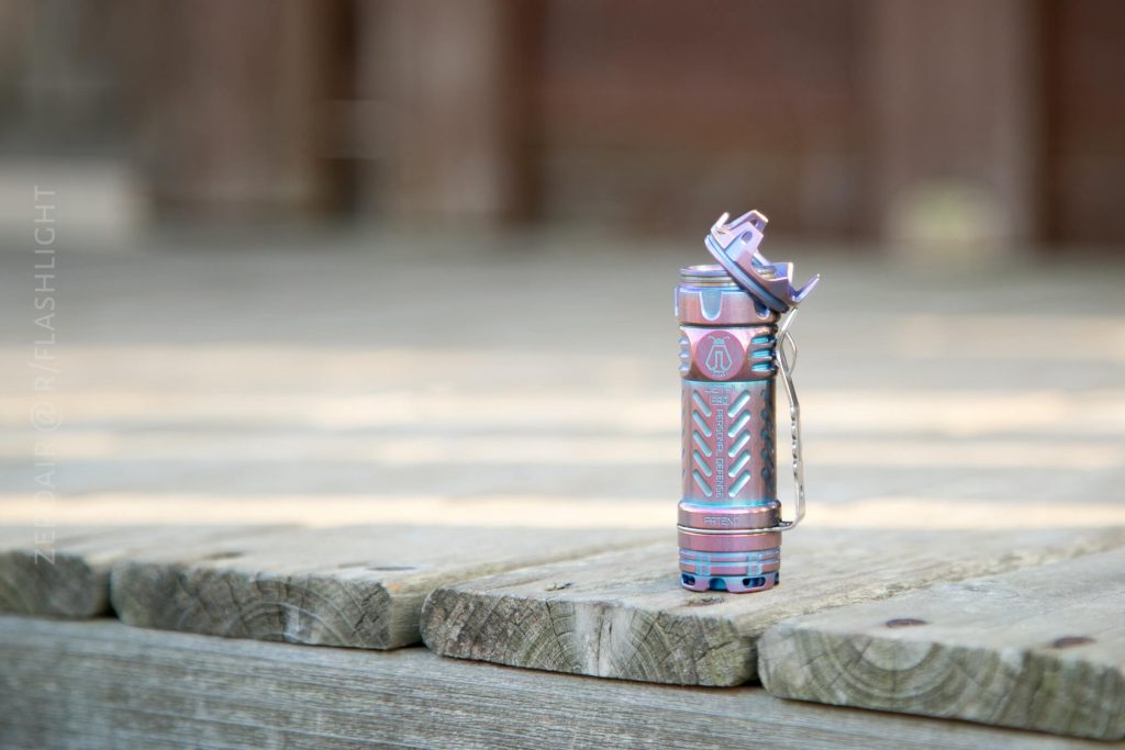 A small, metallic, multicolored flashlight with a pocket clip stands upright on a weathered wooden surface with a blurred background.