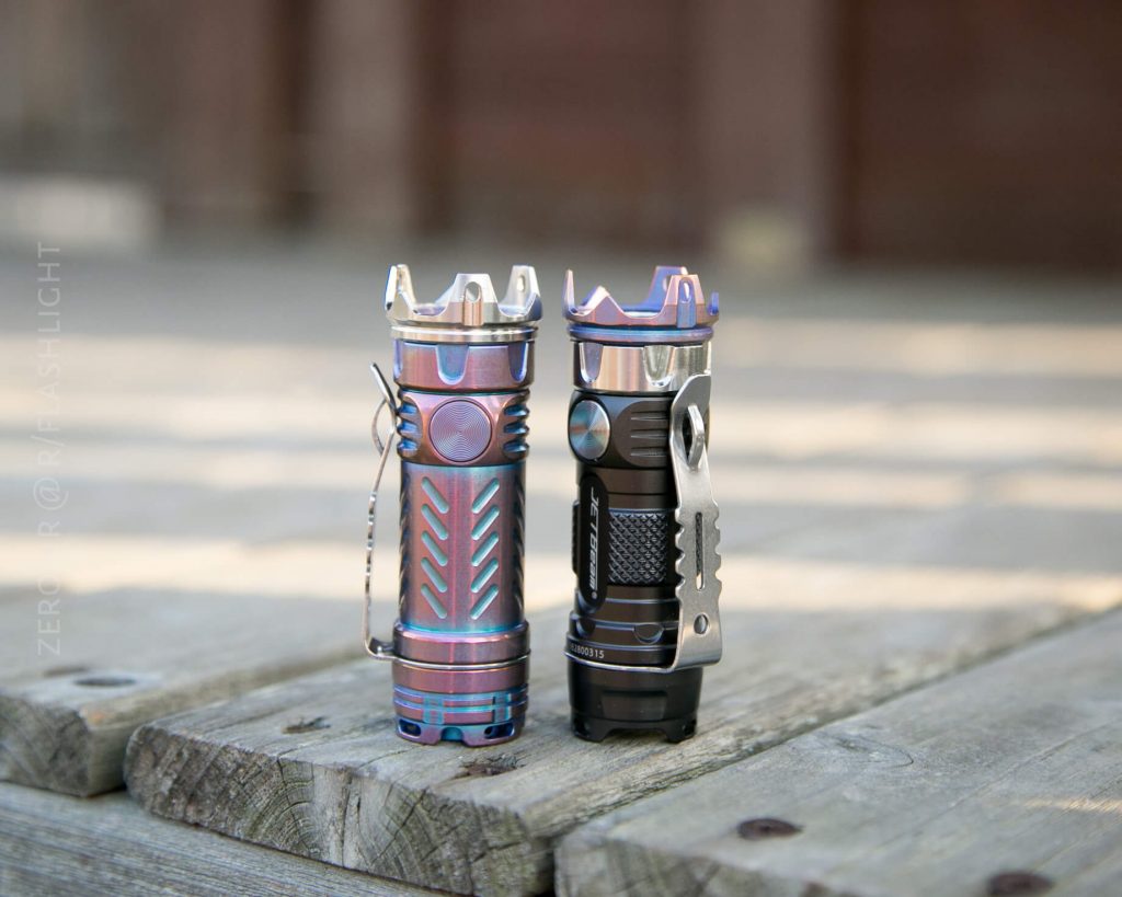 Two compact flashlights stand upright on a wooden surface outdoors. The left flashlight has a metallic, iridescent finish with blue and purple hues, while the right one is mostly black with silver and purple accents.